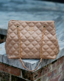 A quilted beige leather tote bag with gold-tone chain straps and a gold monogram logo, resting on a stone ledge above a brick wall.