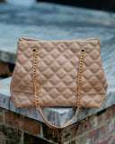A beige quilted leather handbag with gold chain straps, resting on a stone ledge over a brick wall.