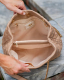 Close-up of a beige quilted handbag being opened by two hands, showing the interior lining, zip pocket, and a gold chain strap on a stone surface.