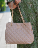 A quilted light beige tote bag with gold chain straps and a gold monogram logo, carried by a person wearing a green patterned dress.