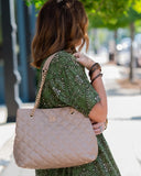 A woman wearing a green patterned dress is shown from behind, carrying a large quilted beige handbag with a gold chain strap over her shoulder in an outdoor street setting.