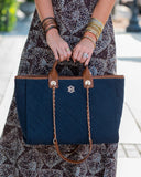 A navy blue quilted tote bag with brown leather handles and trim, held by a person wearing a patterned dress and stacked bangles. The bag features a gold-tone metal logo and a gold chain strap.
