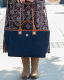 A navy blue quilted tote bag with brown leather handles and a gold-tone logo, held by a person wearing a brown patterned dress and tan boots.