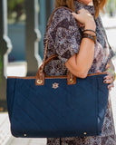 A navy blue quilted tote bag with brown leather handles and a gold monogram logo, carried on the shoulder of a person wearing a patterned dress.