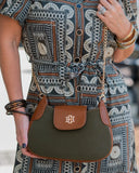 A woman wearing a brown, black, and white patterned dress carries an olive green and brown crossbody bag with a gold chain strap and a gold monogram logo on the front flap.