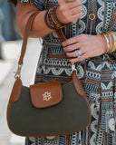 A close-up of a person holding a green and brown shoulder bag with a gold monogram logo. The person is wearing a patterned dress and multiple bracelets.