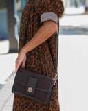 A person wearing a brown leopard-print dress carries a dark brown crossbody bag with a gold monogram logo on a sunny outdoor walkway.