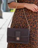 A dark brown leather shoulder bag with a gold-tone interlocking monogram logo on the front flap, worn crossbody over a brown and black leopard print dress.