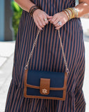 A navy blue and brown striped dress with a matching navy and brown shoulder bag featuring a gold-tone 'T' logo clasp, held by a model wearing gold bangles.