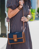 A navy blue and brown crossbody handbag with a gold-tone 'BH' logo clasp, worn by a model in a navy and brown vertically striped dress.