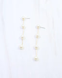 A pair of gold drop earrings with five white pearl beads arranged in a wavy vertical line, displayed on a light white marble background.