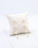 A pair of gold dangle earrings with white faux pearls, displayed on a cream-colored square pillow against a white background.
