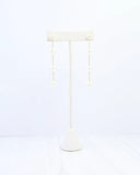 A pair of long drop earrings with small white pearl-like beads on thin gold-tone wires, displayed on a cream jewelry stand against a white background.
