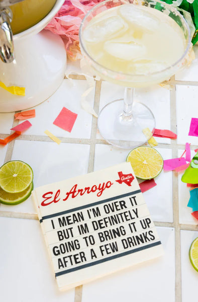 Glass of margarita with lime wedges, colorful straws, and a sign reading 'El Arroyo' on a checkered tablecloth.