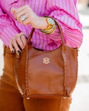 A brown leather handbag with braided trim and a gold interlocking logo, carried by a person wearing a pink striped blouse and brown pants.