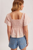 Woman wearing a pink smocked top and denim shorts on a plain background