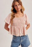 Woman wearing a pink smocked top and denim shorts on a plain background
