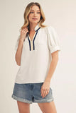 Woman wearing a white blouse with navy accents and denim shorts on a plain background