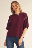 A woman wearing a burgundy knit sweater with white contrast stitching along the shoulders and sleeves, paired with blue jeans, against a plain beige background.