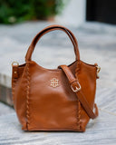A brown leather handbag with top handles, a detachable shoulder strap, decorative whipstitching along the sides, and a gold-tone monogram logo on the front, photographed outdoors on a stone surface.