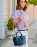 Woman in a lavender blouse holding a navy handbag on a sidewalk.