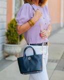 Woman holding a navy blue handbag with gold accents, wearing a light purple blouse and white pants.